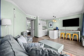 A modern living room with a sofa, a coffee table, a TV, and a kitchen area with a dining table and stools. - Beach View Retreat,KING,Pools,M149 (Myrtle Beach)