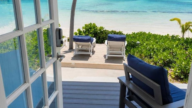 A view from a balcony with a wooden deck, two lounge chairs with blue cushions, and a clear view of a turquoise ocean and beach. - Bella Vista Breeze - Beachfront Oasis awaits your arrival. (George Town)