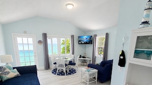 A spacious living area with a sofa, a TV, a dining table, and a view of the sea. - Bella Vista Breeze - Beachfront Oasis awaits your arrival. (George Town)