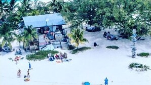 A beach with two boats docked, a building with a blue roof, and people engaging in various activities. - Bella Vista Breeze - Beachfront Oasis awaits your arrival. (George Town)