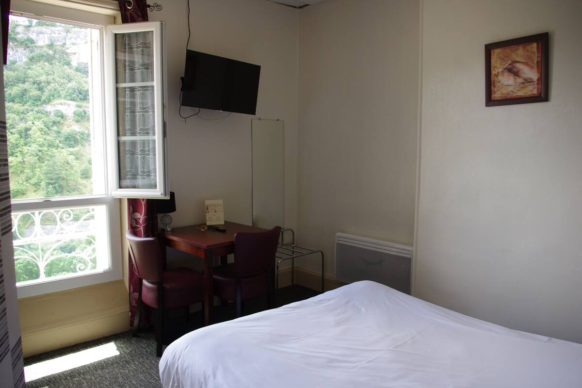 Habitación de hotel con cama, escritorio con silla, televisor en la pared y ventana con cortinas.