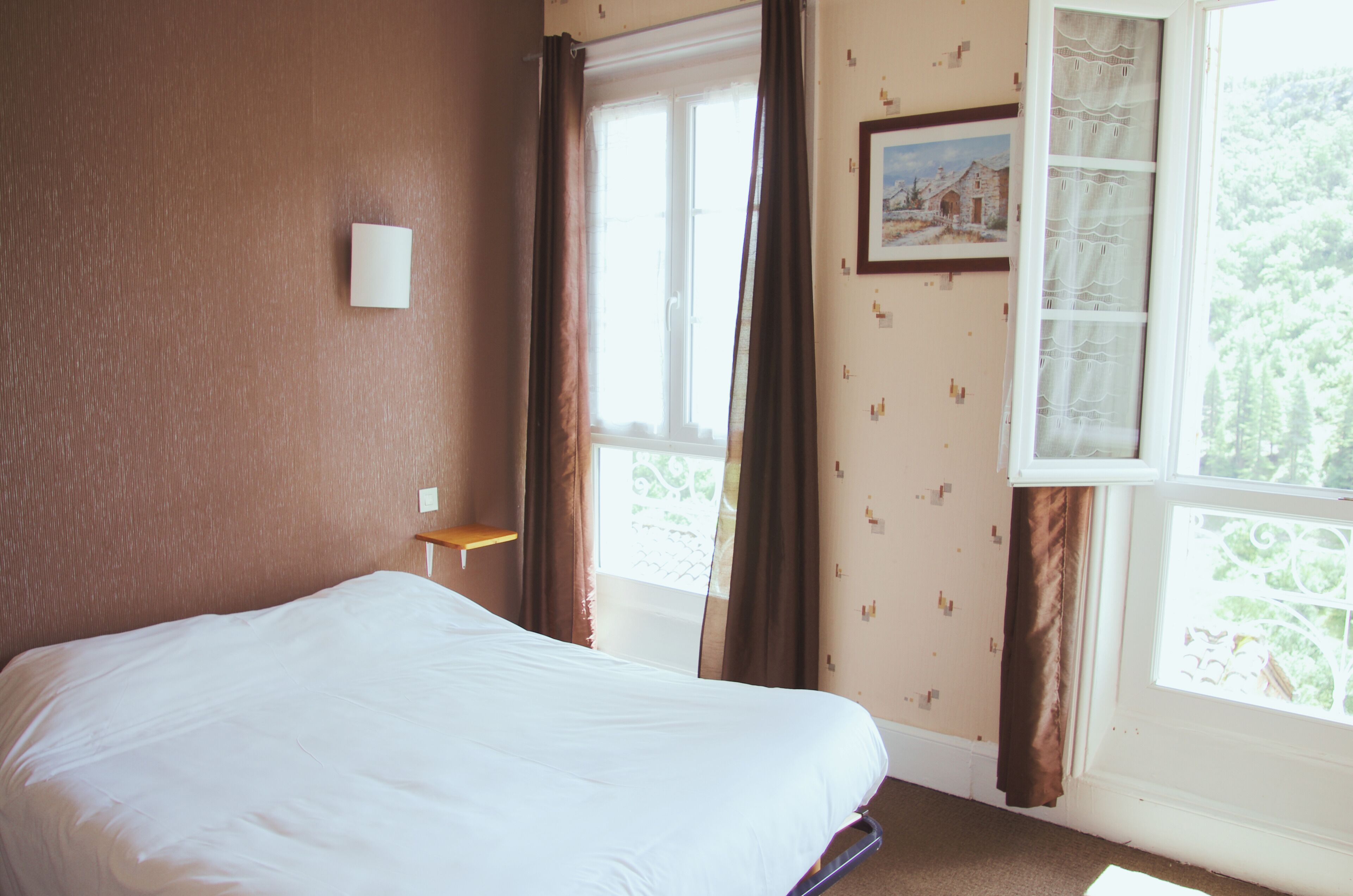 A hotel room with a large bed, a window with curtains, a small table, and a wall-mounted light.