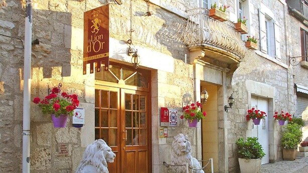 Un bâtiment en pierre avec une porte en bois et une statue de lion devant. Des pots de fleurs sont placés sur les rebords des fenêtres et une enseigne est accrochée au-dessus de la porte.