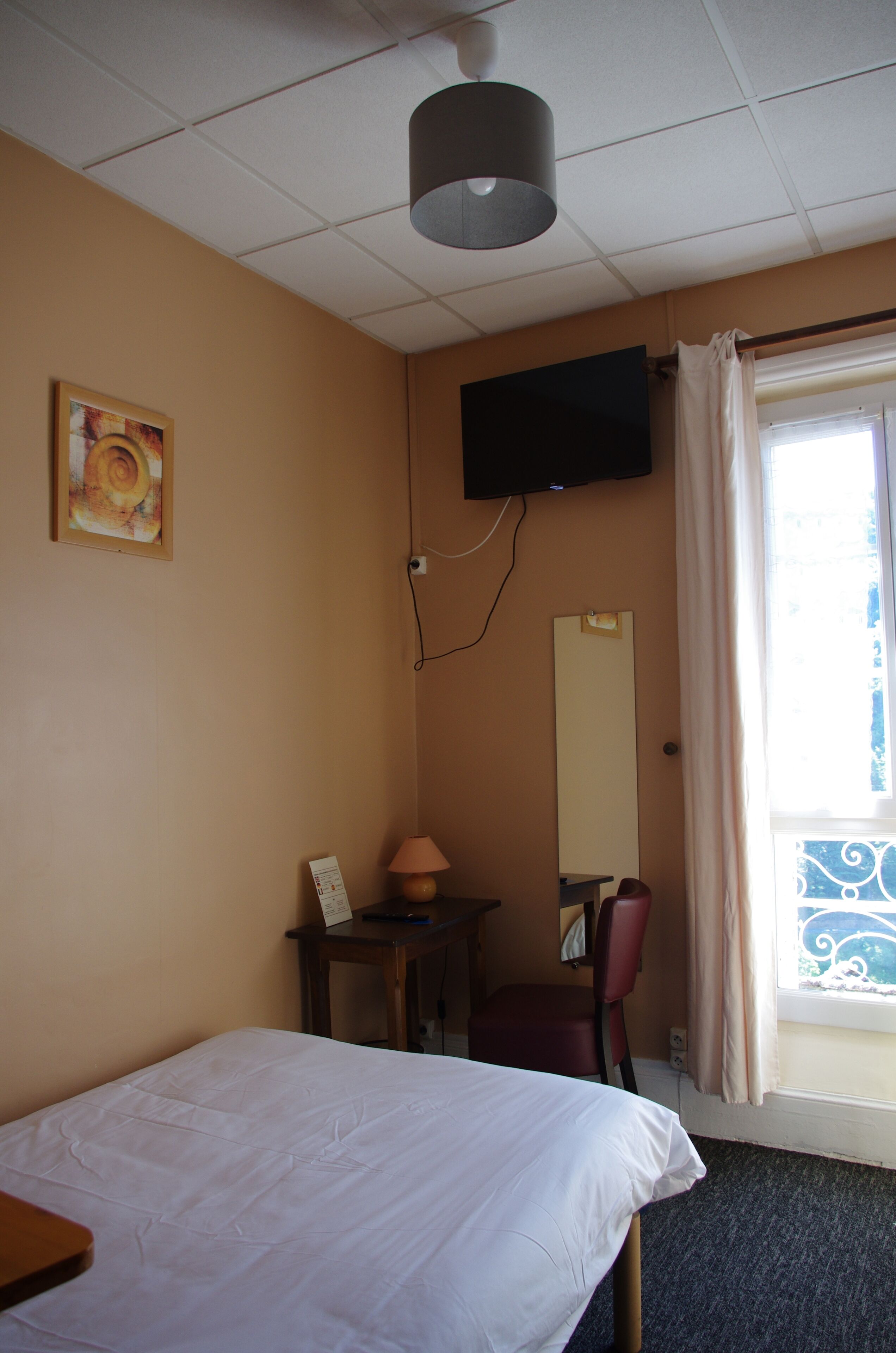 Une chambre d’hôtel comprenant un lit, un bureau, une chaise, un téléviseur fixé au mur, une fenêtre avec des rideaux et un tableau au mur.