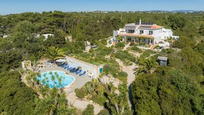 A large white villa with a swimming pool and lounge chairs, surrounded by dense greenery and a clear blue sky.