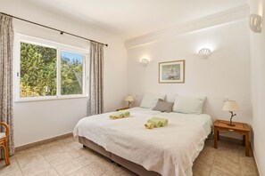 A bedroom with a bed, two bedside lamps, a window with curtains, a wall-mounted picture, and a small bedside table.