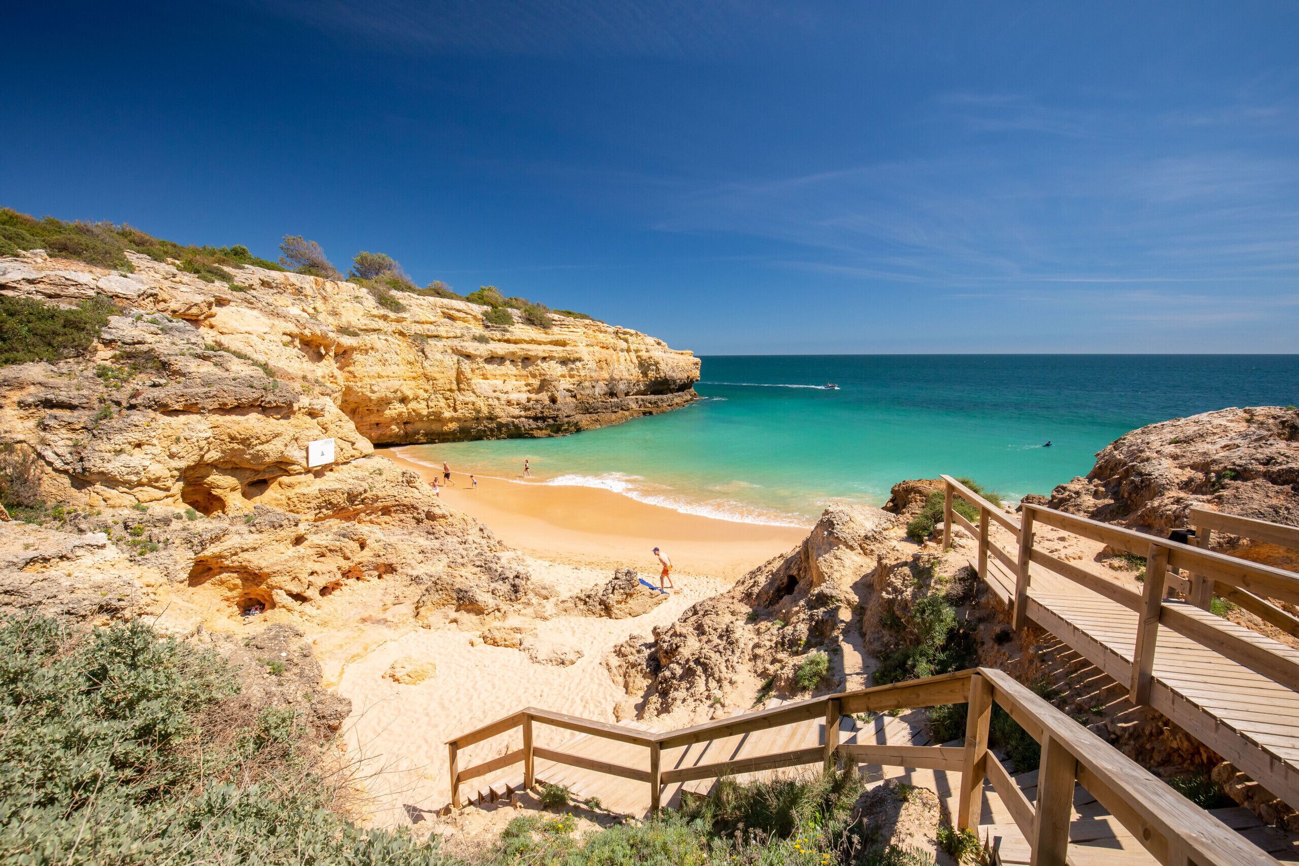 A beach with a wooden staircase leading down to the shore, clear blue water, and rocky cliffs.