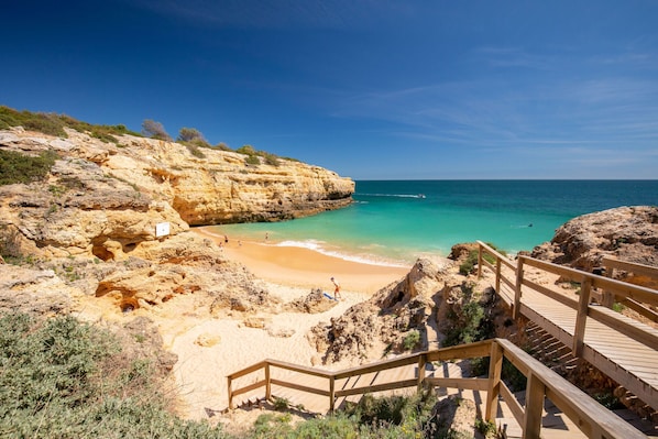 A beach with a wooden staircase leading down to the shore, clear blue water, and rocky cliffs.