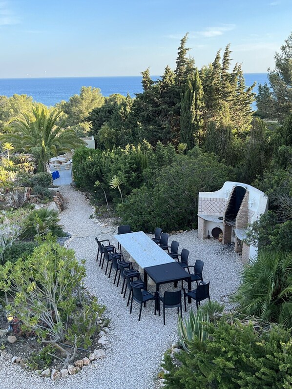 An outdoor patio with a long table and chairs, surrounded by gravel and greenery, with a barbecue grill and a view of the sea.