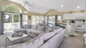 A spacious living room with a large sectional sofa, a dining area with a table and chairs, a kitchen with white cabinets and appliances, and a ceiling fan. - Beach House (Corolla)
