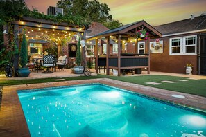 A backyard with a swimming pool, patio furniture, a covered seating area, and a brick house.