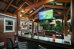 An outdoor bar area with a television displaying a cartoon, a grill, and seating.