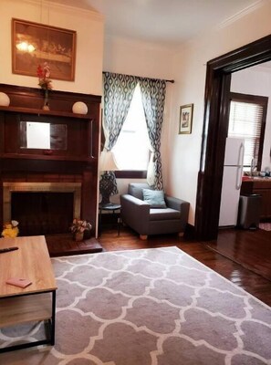 Cozy living room with a fireplace, comfortable armchair, and dining table, featuring a patterned rug and framed picture.