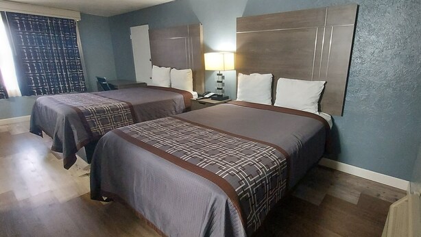 A hotel room with two beds, a nightstand, a lamp, and a window with curtains. - HOTEL BLUE (DeLand)