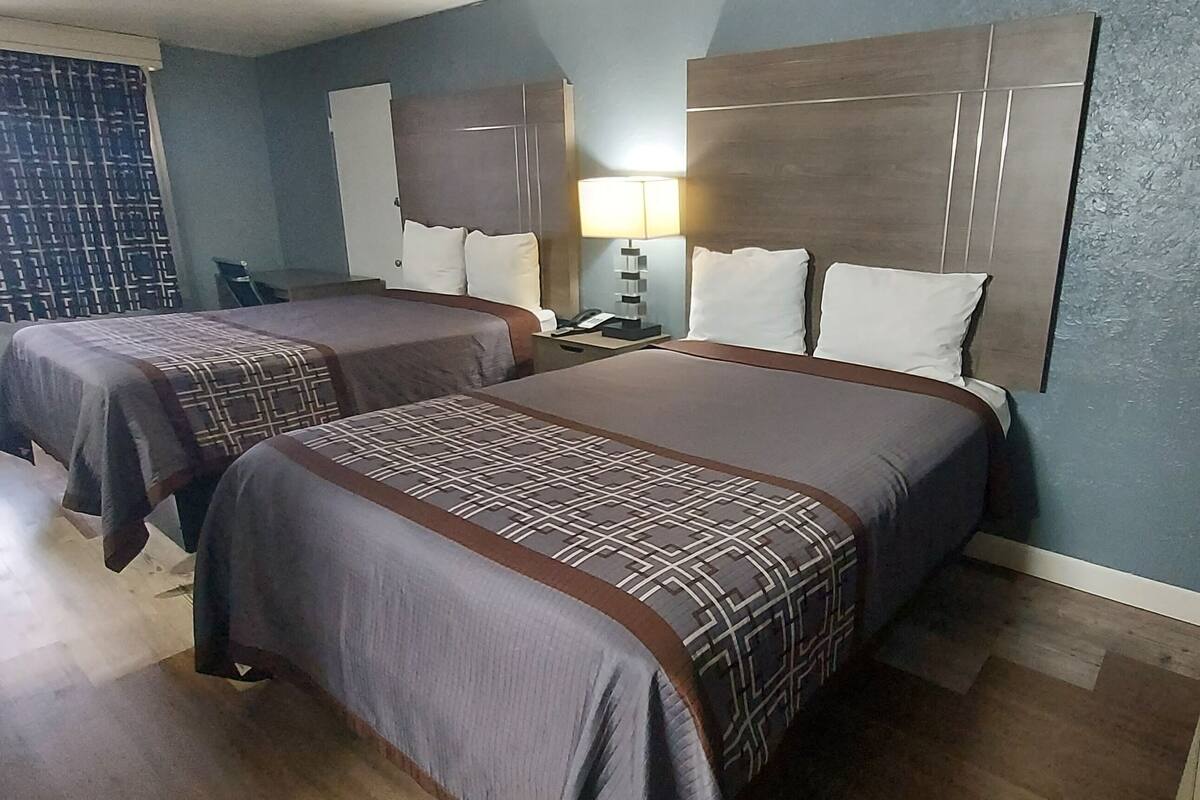 A hotel room with two beds, a nightstand, a lamp, and a window with curtains.
