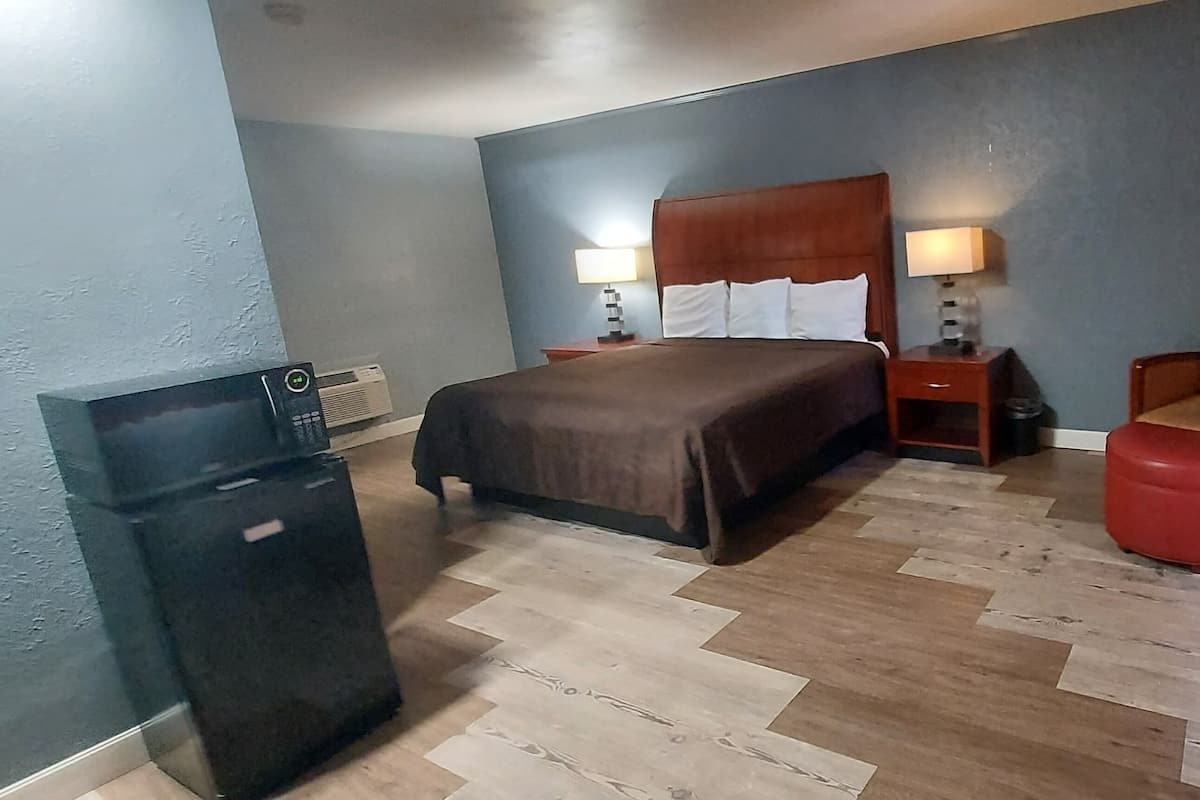 A hotel room with a bed, bedside tables, a chair, a red ottoman, and a refrigerator.