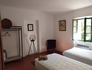 A bedroom with two beds, a standing lamp, a chair, a coat rack, and a window with green shutters.