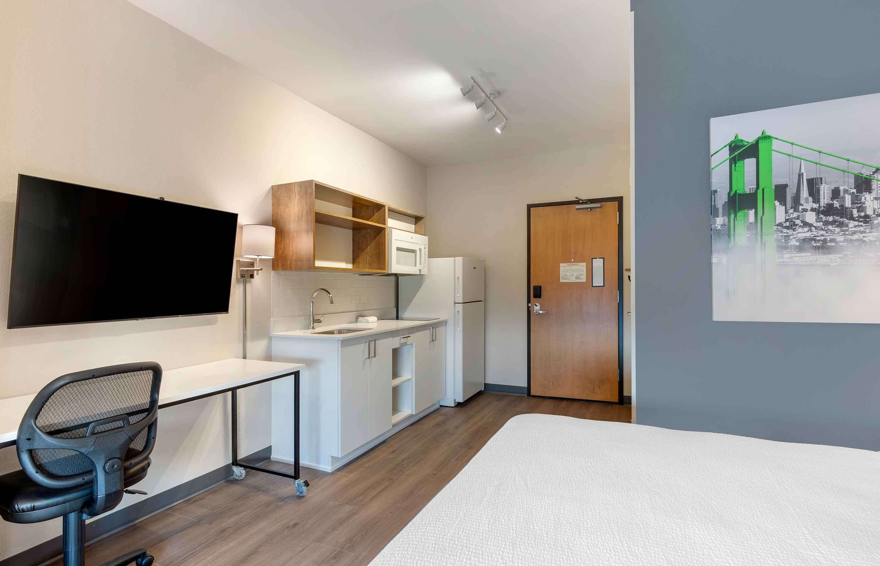 Een kleine hotelkamer met een kitchenette, een flatscreen televisie, een bureau met stoel, een bed en een deur.