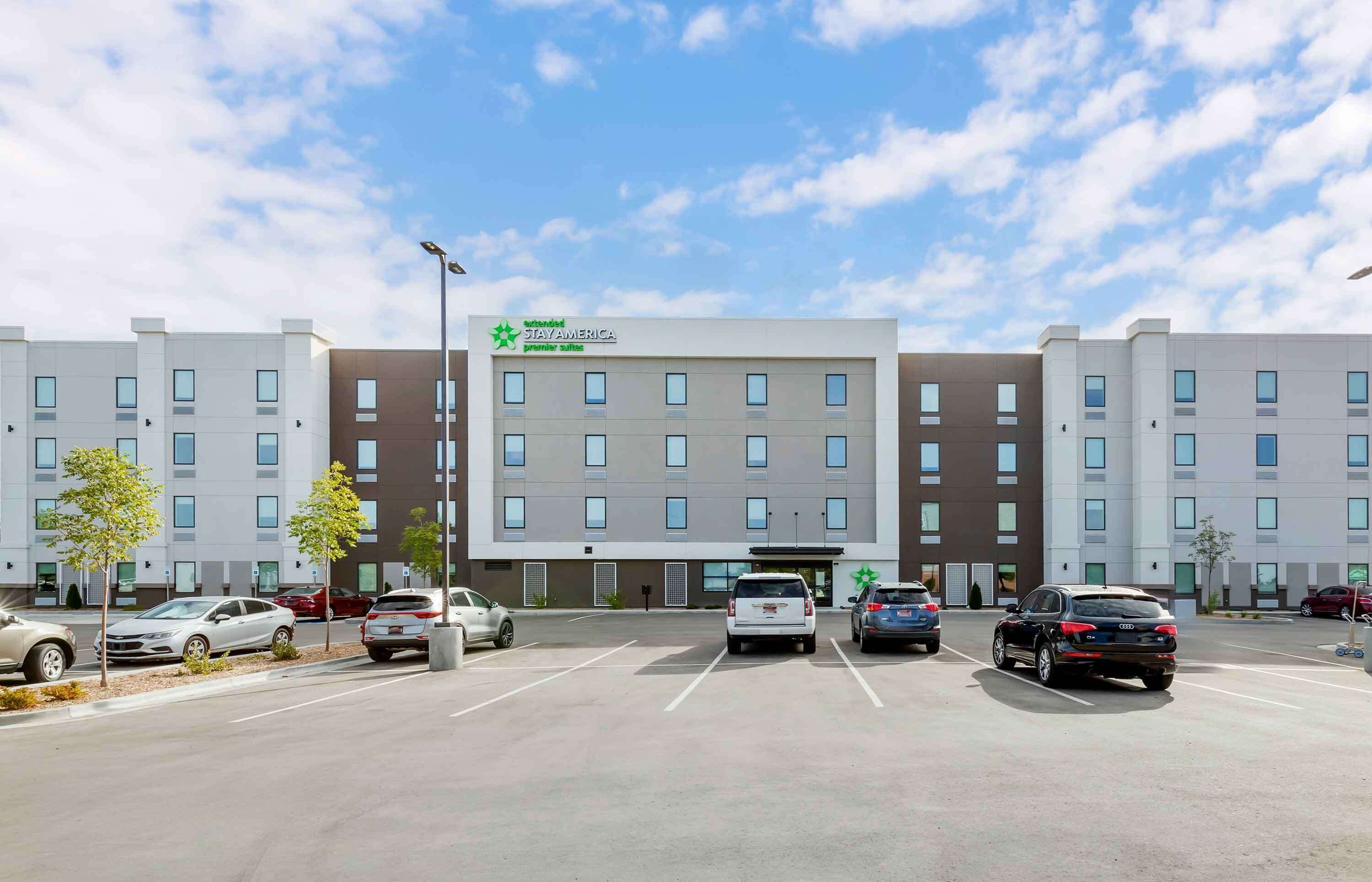 A Stay America hotel with a modern design, featuring a central entrance, multiple parking spaces, and a clear sky.