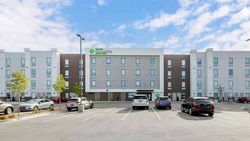 A Stay America hotel with a modern design, featuring a central entrance, multiple parking spaces, and a clear sky.