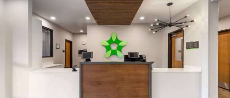 A reception area with a wooden reception desk, a green logo on the wall, and a modern ceiling design.