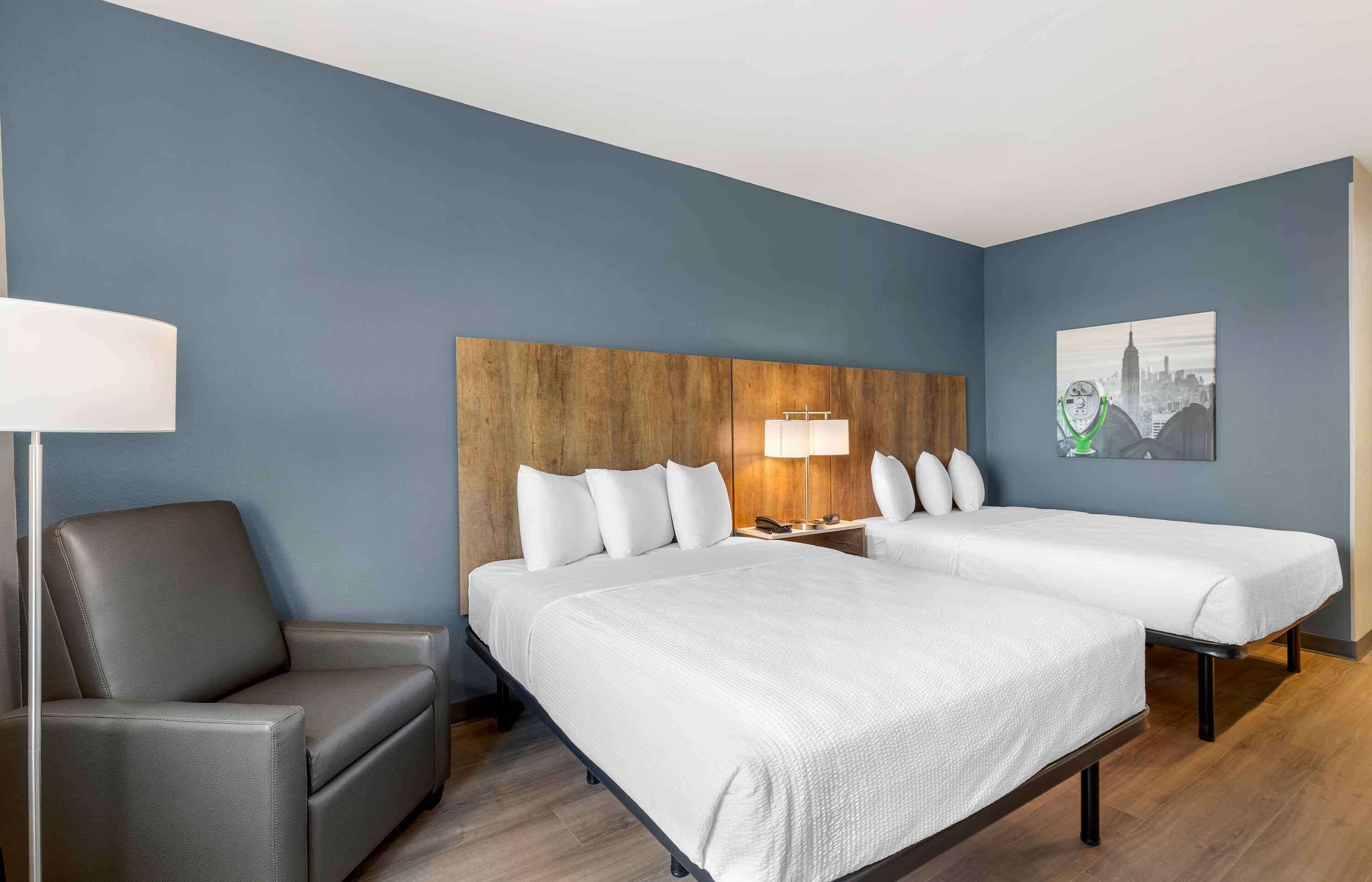 A hotel room with two beds, a sofa, a nightstand with a lamp, and a cityscape painting on the wall.