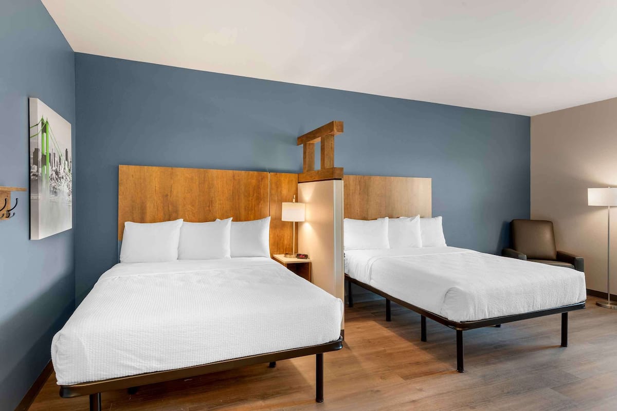A hotel room with two single beds, a wooden headboard, a nightstand with a lamp, a chair, and a floor lamp.