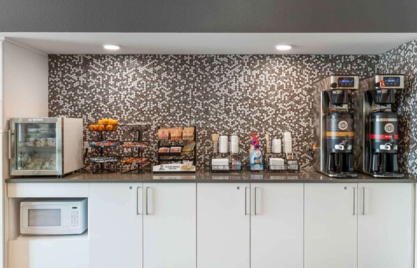 A kitchenette with a microwave, refrigerator, coffee machines, and a variety of food items.