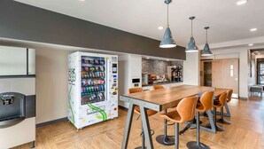 A modern dining area with a vending machine, a long wooden table, and orange chairs. - Extended Stay America Premier Suites - Boise - Meridian (Meridian)