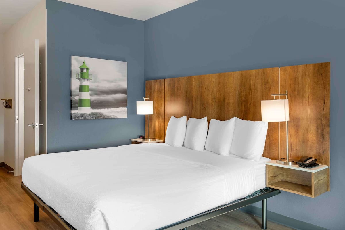 A hotel room with a large bed, wooden headboard, two bedside tables with lamps, a painting of a lighthouse, and a door to the left.