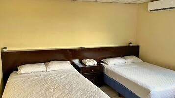Quarto de hotel com duas camas de solteiro, uma pequena mesa de cabeceira com abajur e um ar condicionado na parede.
