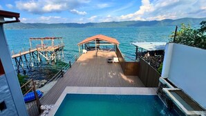 Une terrasse en bois avec une piscine, offrant une vue sur une étendue d’eau et sur des montagnes lointaines.