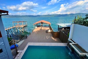 Terraza de madera con piscina, con vista a un cuerpo de agua y montañas distantes.