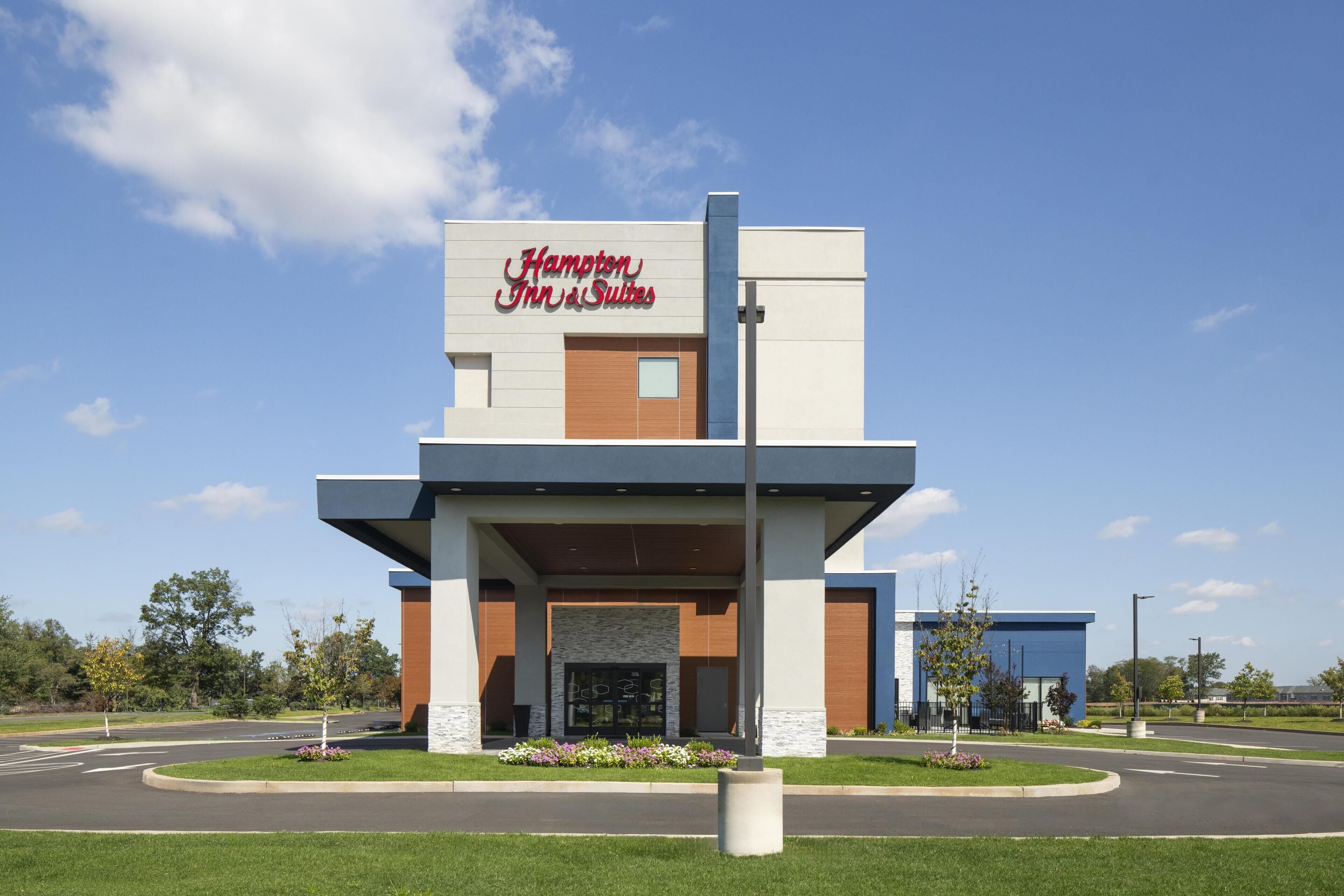 A modern Hampton Inn & Suites with a prominent sign, a central entrance, and landscaped surroundings.