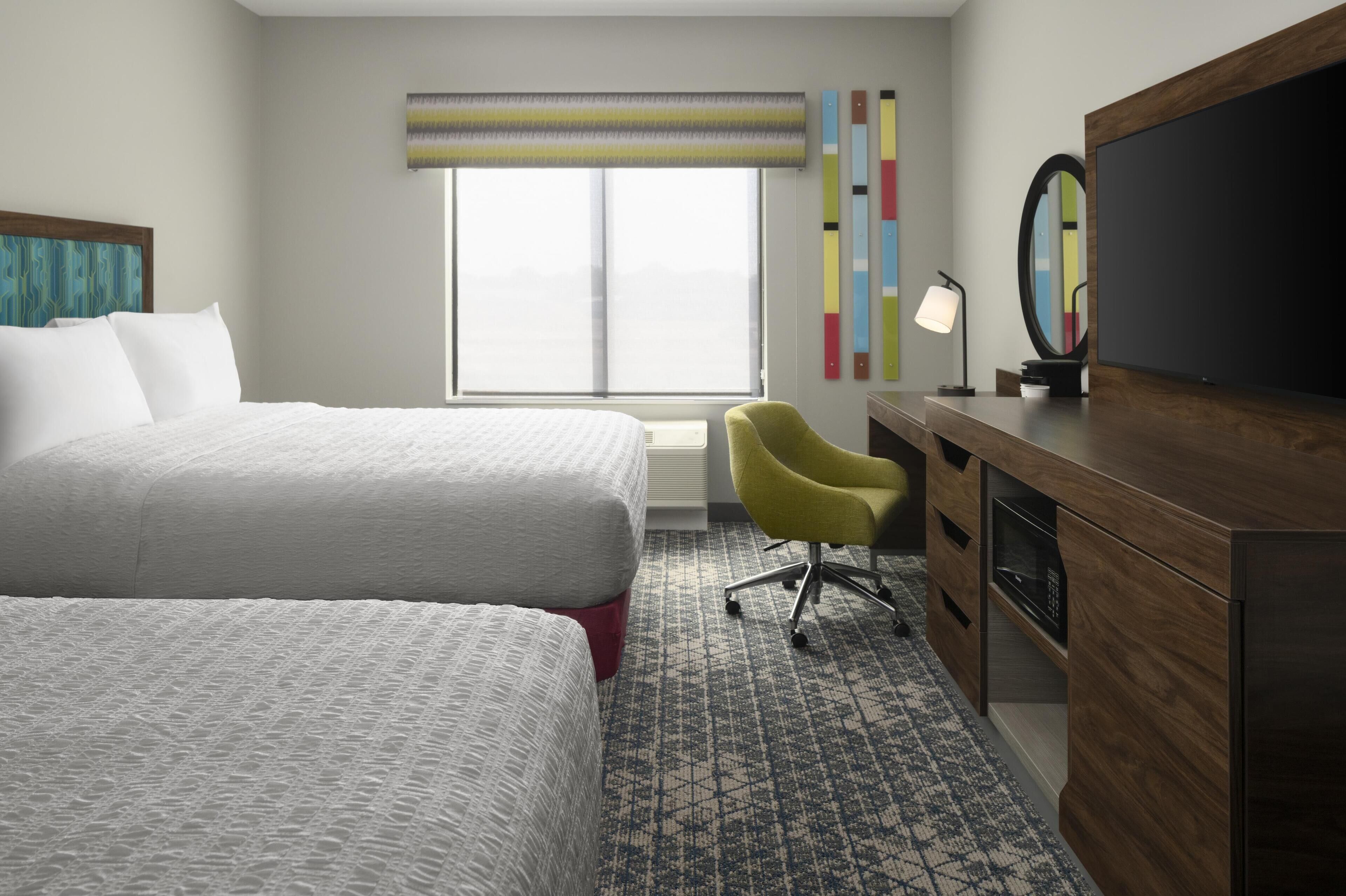 A hotel room with two beds, a desk with a mirror, a chair, and a large window with a view.