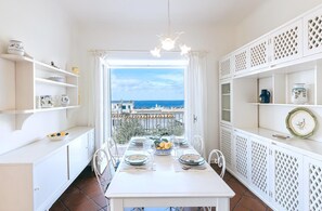 Un comedor con una mesa dispuesta para cuatro, muebles blancos con diseños de celosía y vistas al mar a través de un ventanal.