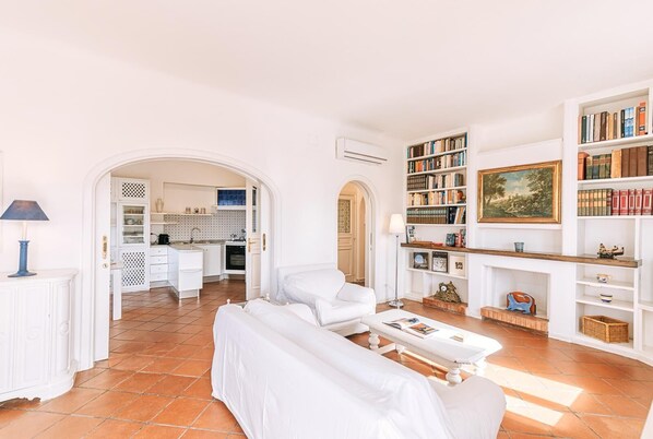 Amplio salón con un sofá blanco, una mesita de centro y una estantería llena de libros. Se ve una cocina a través de una puerta abierta.