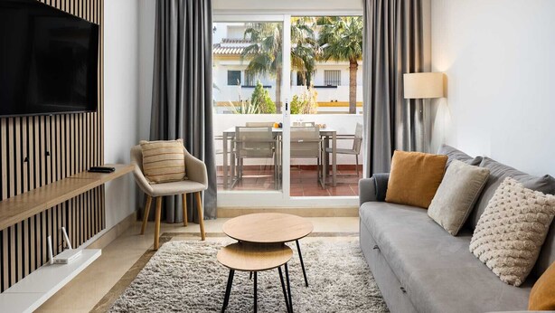 A modern living room with a grey sofa, a wooden TV stand, a round coffee table, and a view of a balcony with outdoor furniture. - Alfresco Stays Calanova Golf Sensations (Mijas Costa)