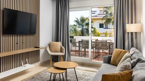 A modern living room with a flat-screen TV mounted on a wooden wall, a sofa with cushions, a round coffee table, and a chair with a striped cushion. - Alfresco Stays Calanova Golf Sensations (Mijas Costa)