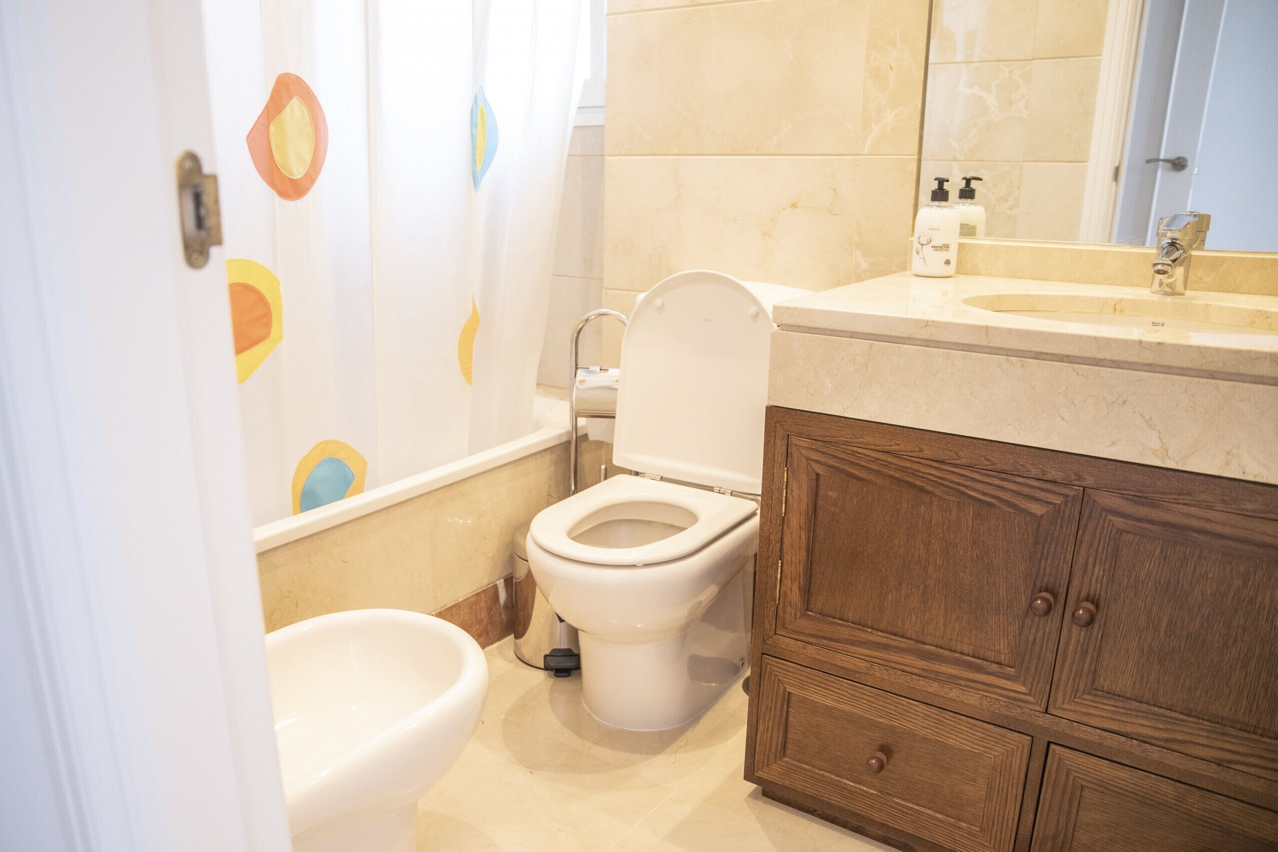 A bathroom with a toilet, a bidet, a wooden cabinet, and a sink with toiletries.
