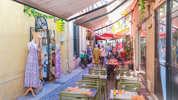 Un café en plein air avec des tables et des chaises, une moto garée devant, et une rue bordée de boutiques et d’une foule de passants.