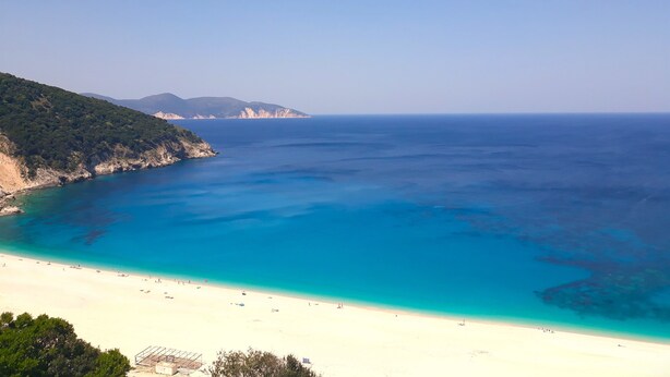 Una playa costera con aguas azul turquesa, orilla arenosa y montañas al fondo.