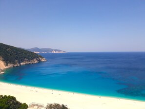 A coastal beach with clear blue waters, sandy shore, and a mountainous backdrop.