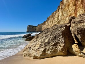A coastal landscape with a sandy beach, large rock formations, and a cliff with a small hill on top.