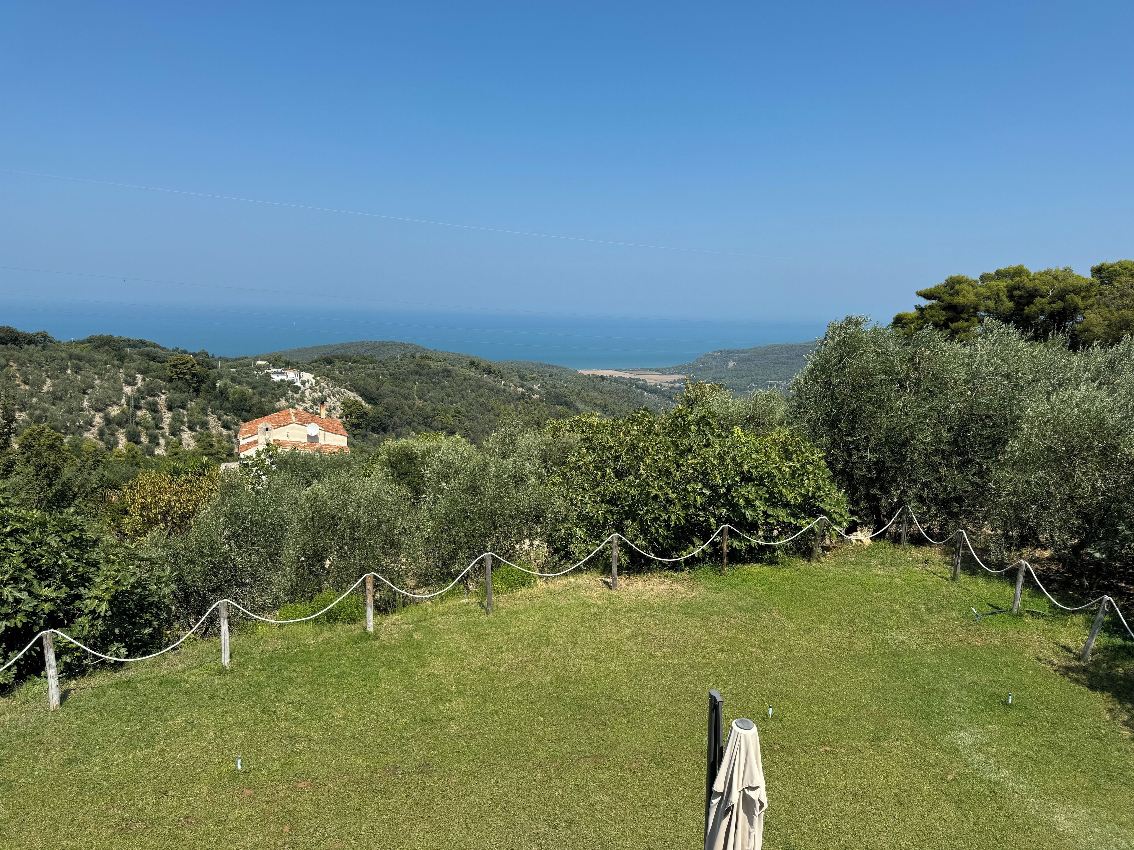 Una zona erbosa con una recinzione, un edificio con tetto rosso e vista sul mare e sulle colline sullo sfondo.