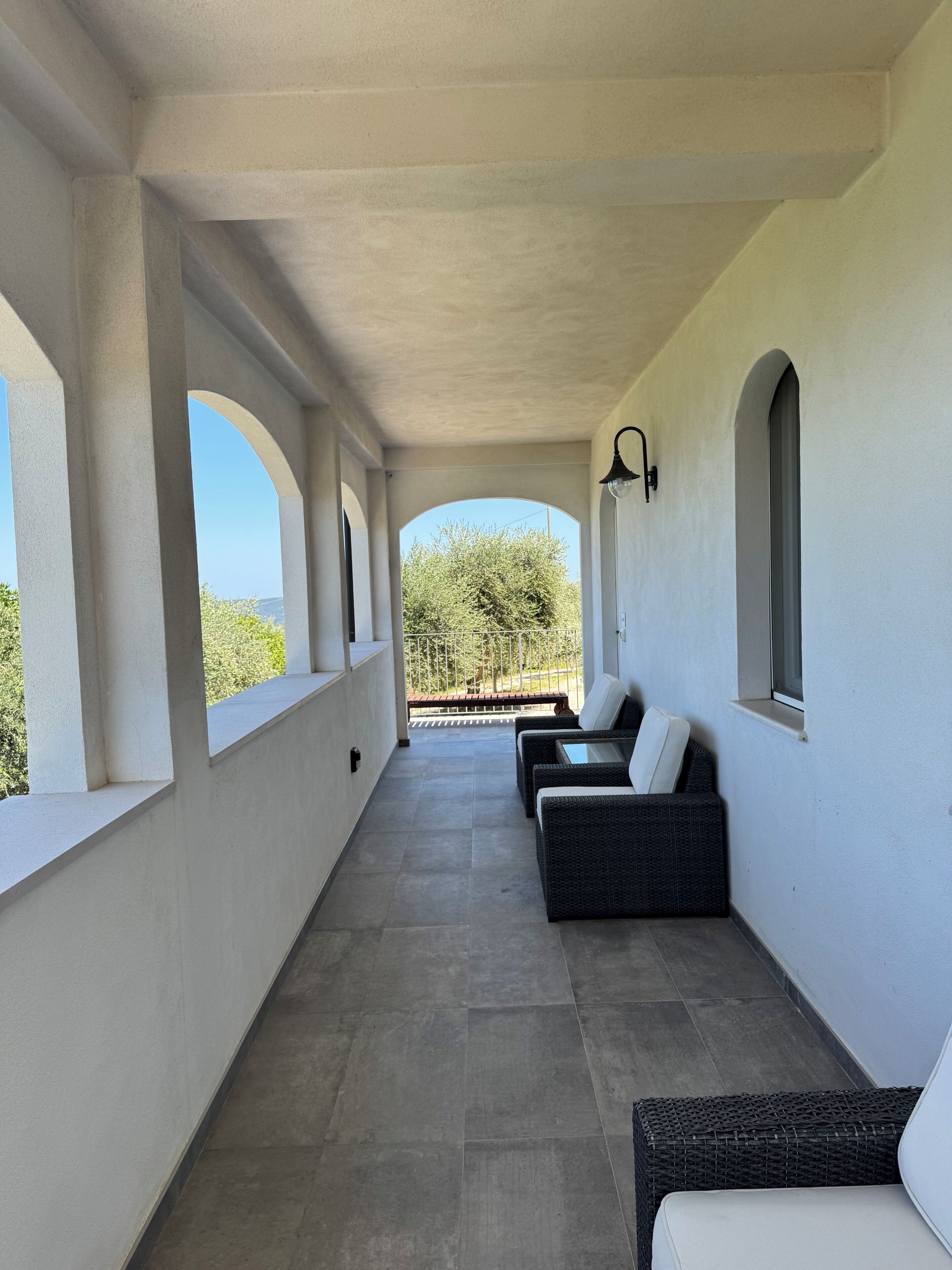 A covered outdoor area with a row of wicker sofas and a bench, large windows, and a view of trees and hills.