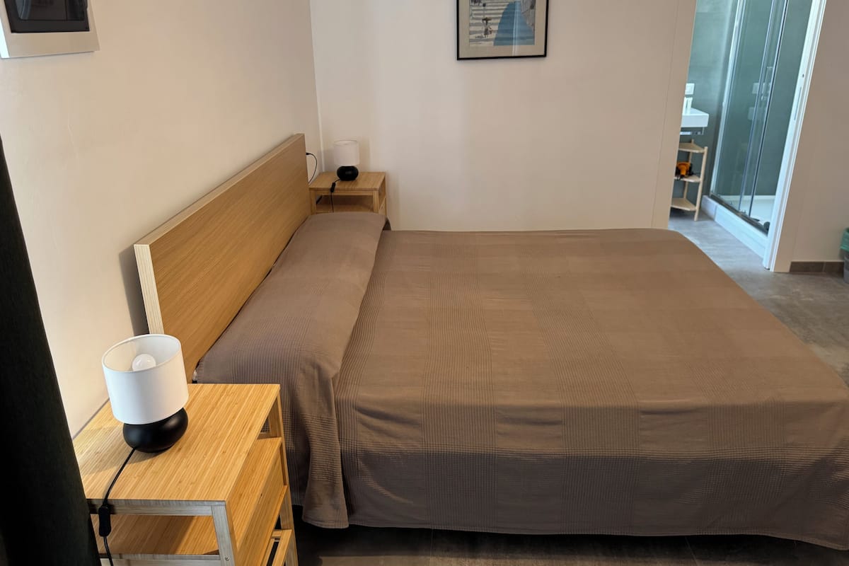 Ein Hotelzimmer mit einem Bett, einem Nachttisch mit Lampe, einem kleinen Tisch ebenfalls mit Lampe, einem Bild an der Wand und einem Fenster mit Blick auf Gebäude.