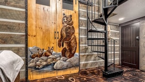 A wooden cabin interior with a bear mural, a spiral staircase, and a stone fireplace.