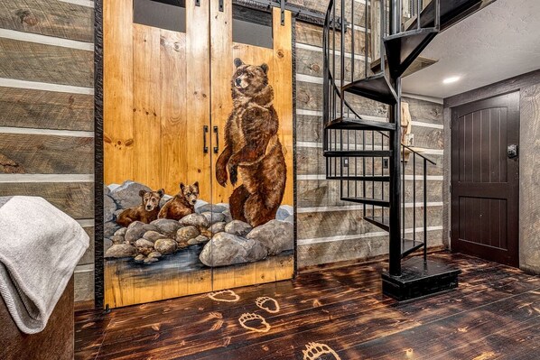 Interior de una cabaña de madera con un mural de un oso, una escalera de caracol y una chimenea de piedra.