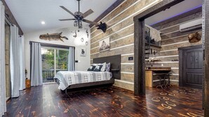 A bedroom with a large bed, wooden flooring, a ceiling fan, and a view of the outdoors.